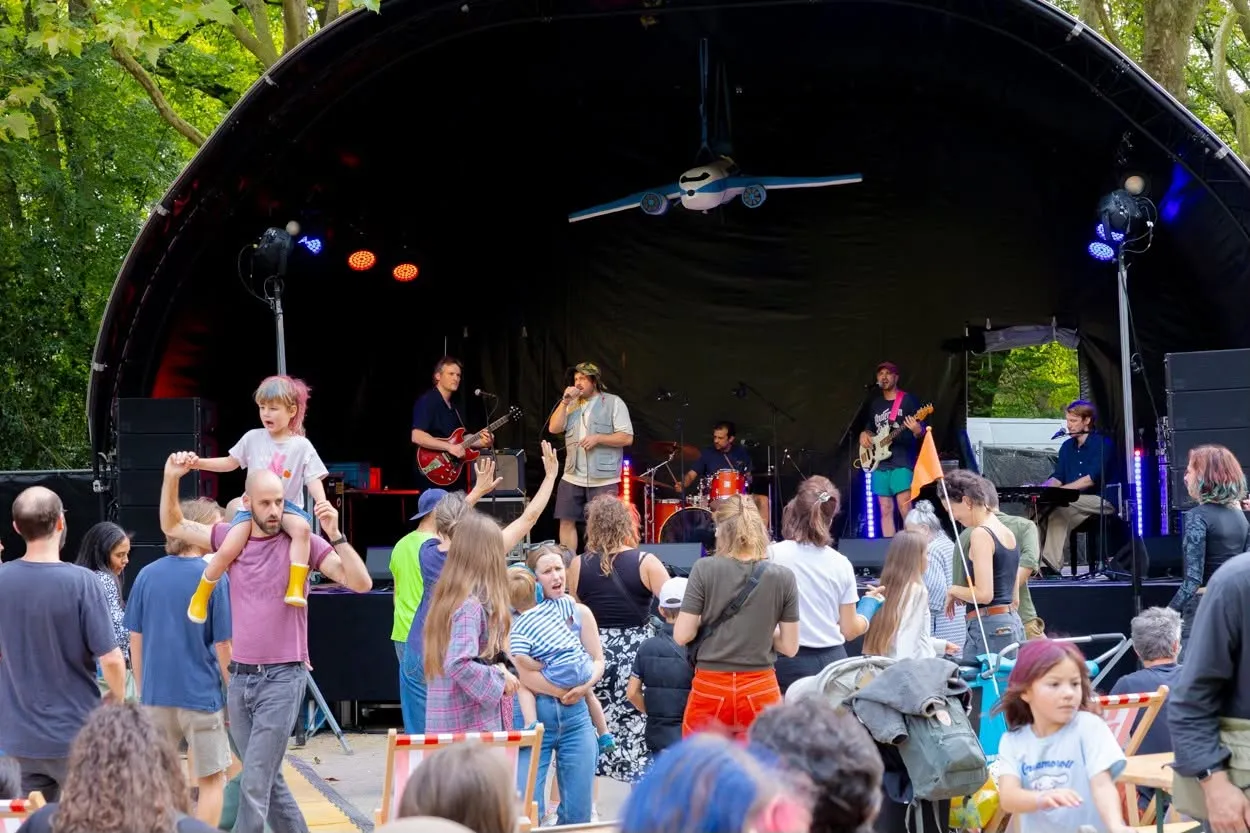 Publiek danst voor het podium, een kind zit op de schouders van een volwassene, het vliegtuig hangt boven de band.