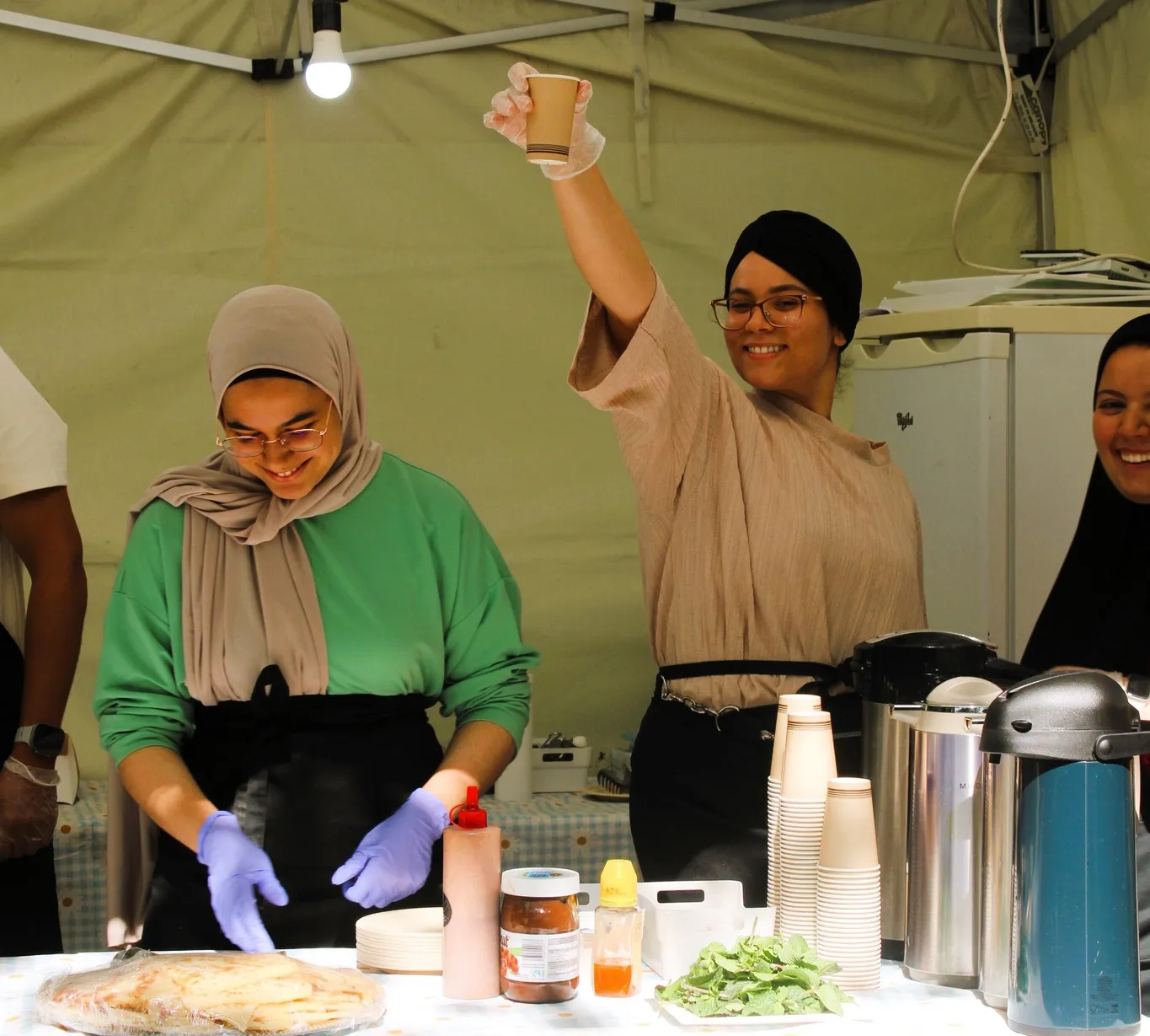 Een eetstand onder een tent op Plazey: twee vrouwen bereiden en serveren eten, één heft een beker op.