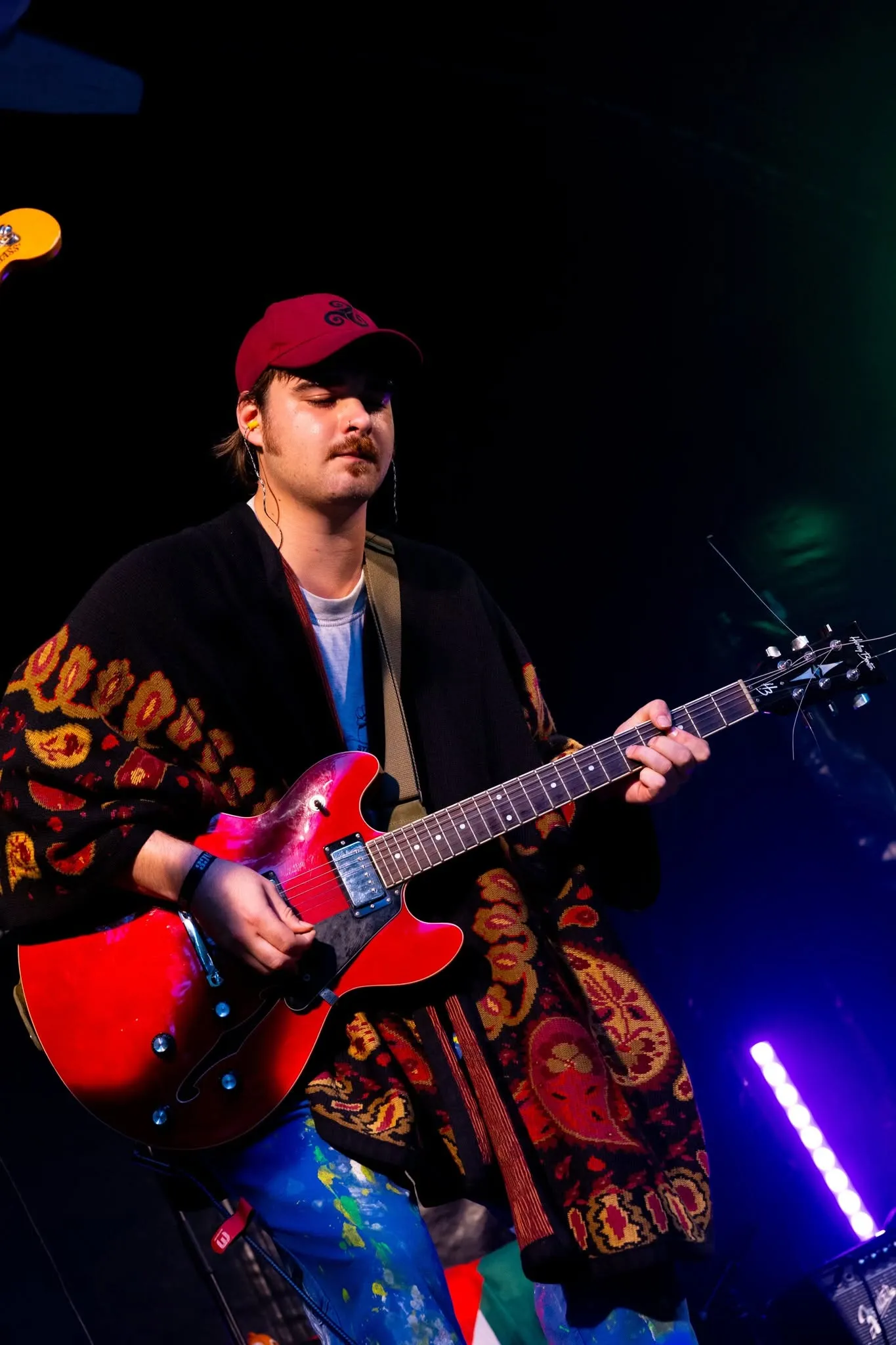 Een gitarist in een gebloemd vest speelt op een rode gitaar, met een pet op.