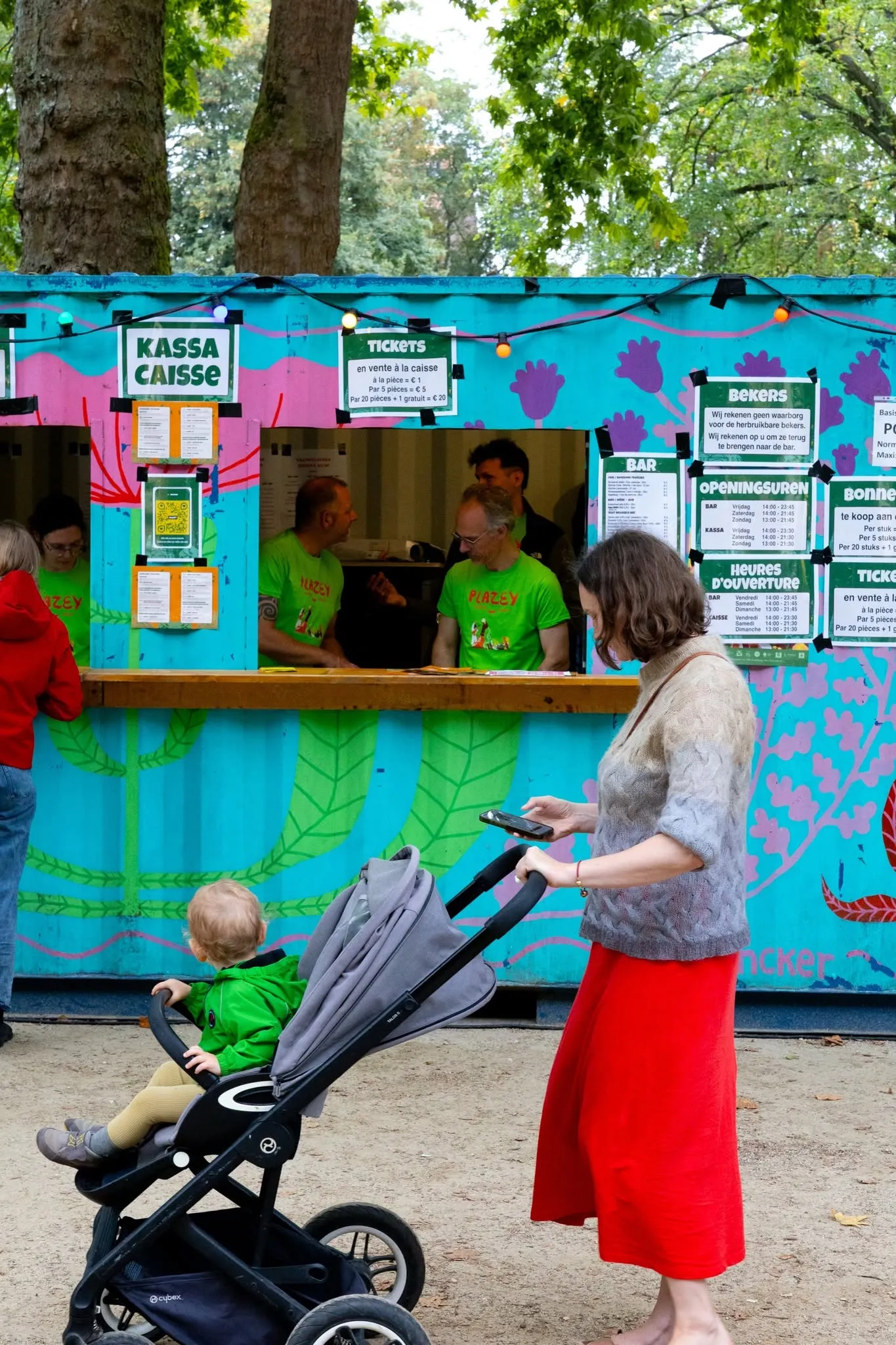 Bezoekers aan de blauwgeschilderde kassa. Medewerkers in groene t-shirts bedienen, een vrouw wacht met een buggy.