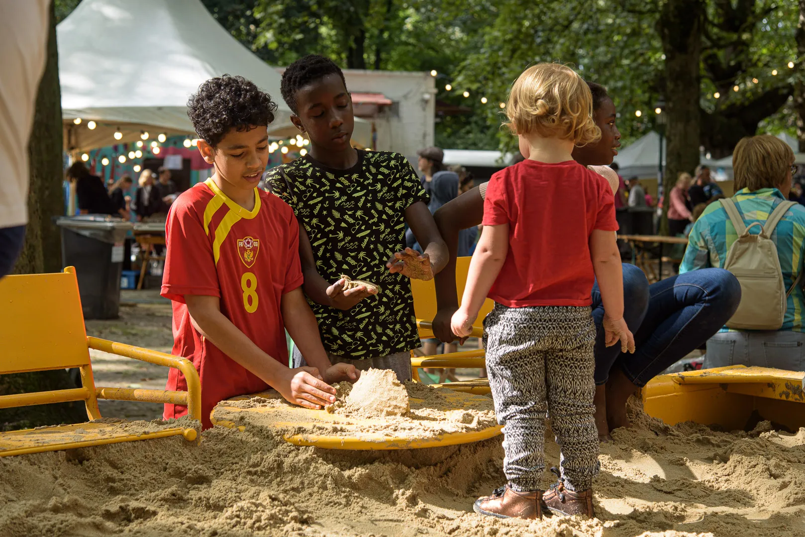 Kinderen van verschillende leeftijden spelen samen in een zandbak op Plazey, met festivalkramen en lichtjes op de achtergrond.