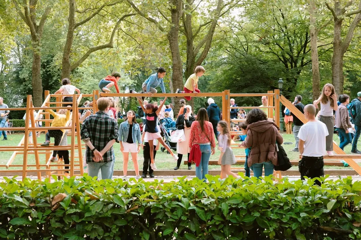 Kinderen klimmen over een houten klimtoestel in het park, ouders kijken toe vanaf het gazon.