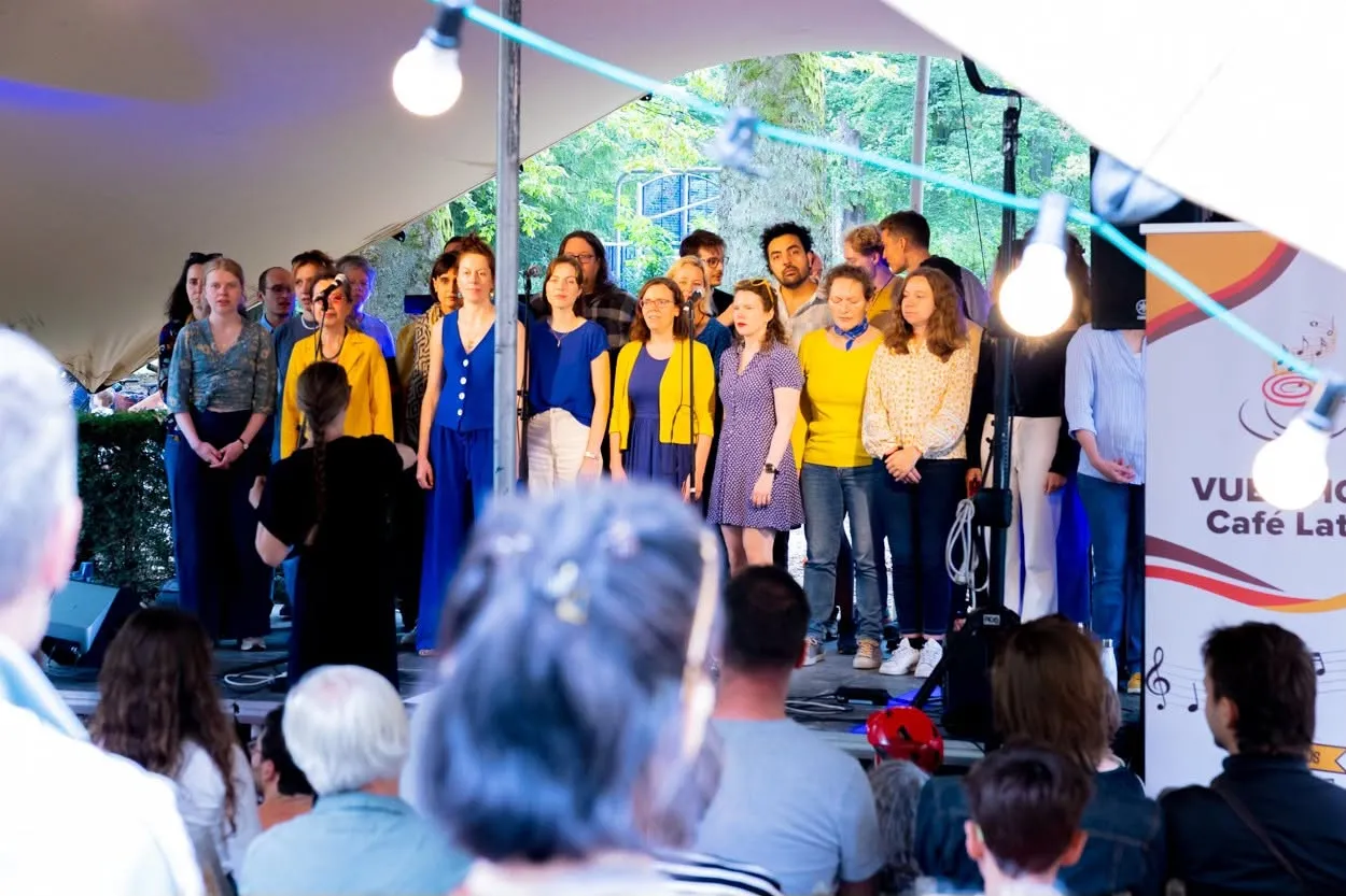 Een koor in blauw en geel zingt onder de tent, met een banner van Café Latino op de achtergrond.