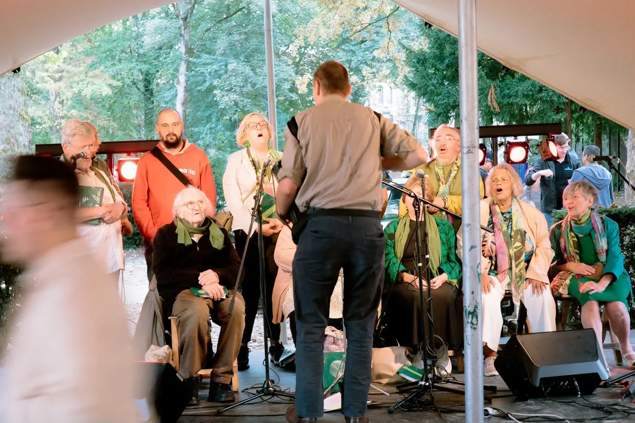 Een koor van jong en oud zingt onder een tent in het park, begeleid door een dirigent.