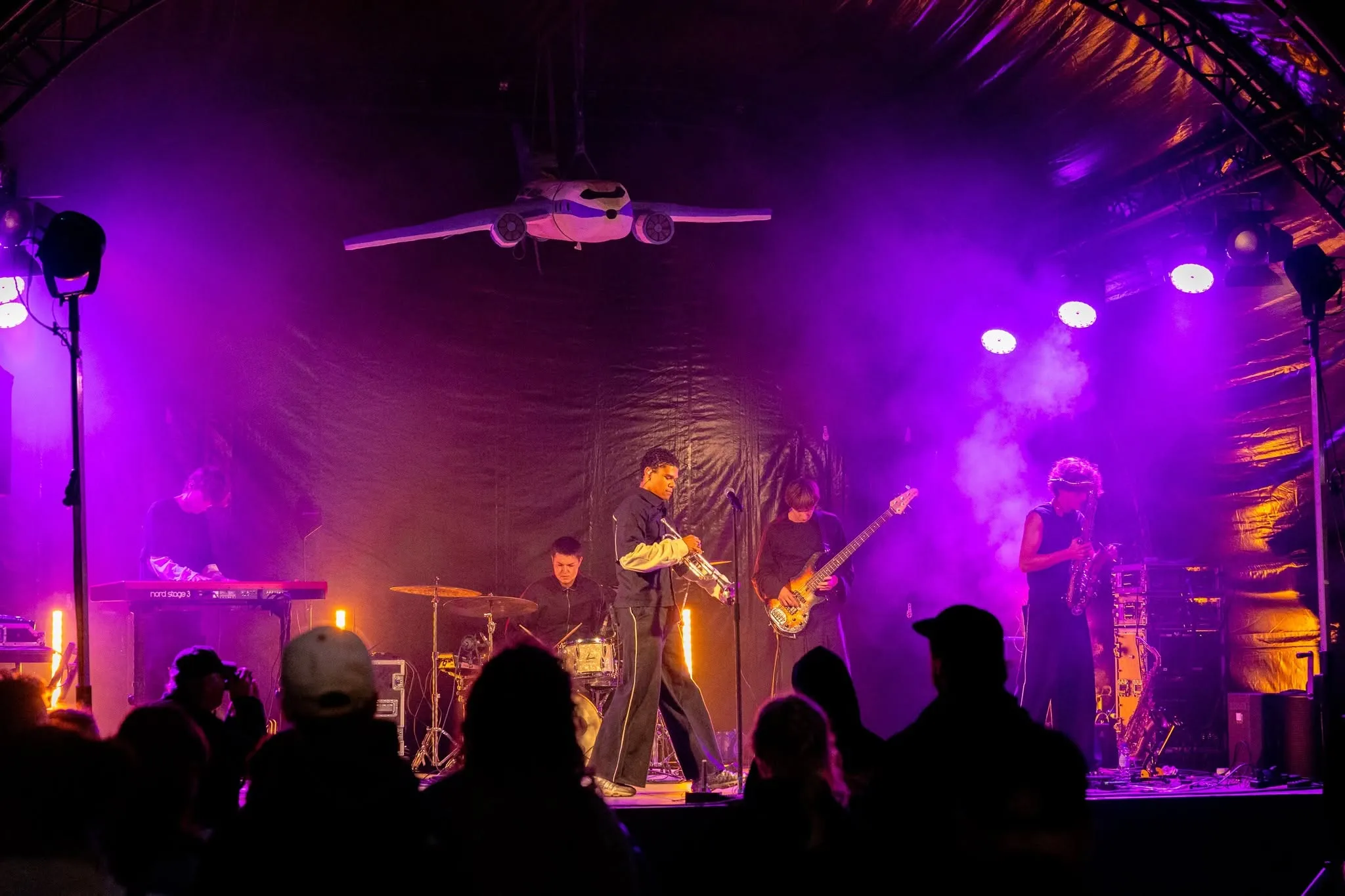 Een band speelt in paars licht onder het vliegtuig dat boven het podium hangt. Silhouetten van het publiek op de voorgrond.