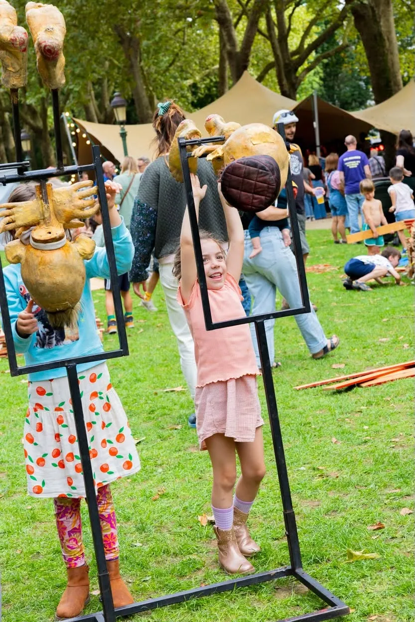 Twee meisjes tillen gouden poppenkoppen op zwarte frames omhoog, lachend in het park.