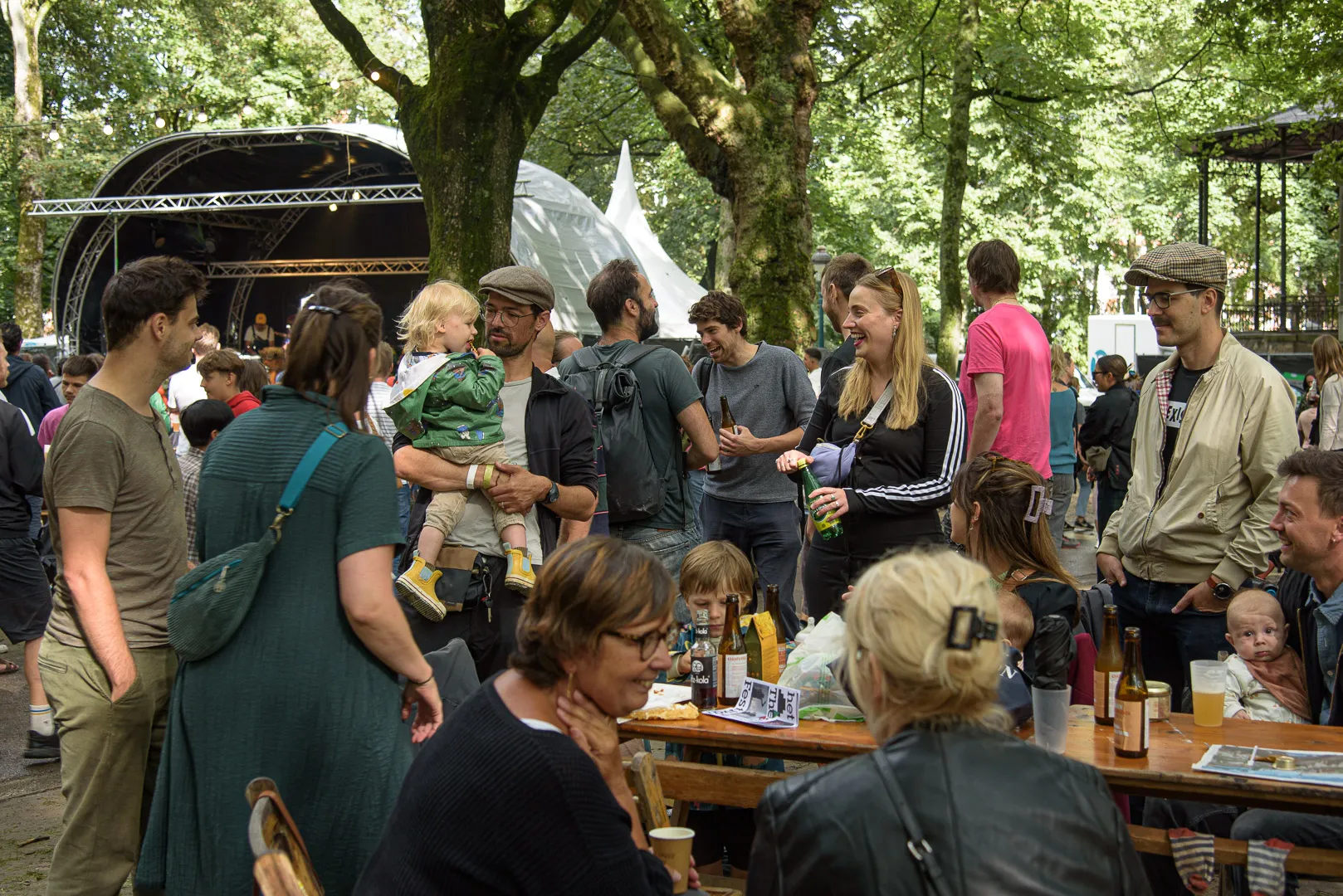 Festivalgangers aan picknicktafels bij het podium, gezinnen, slingers in de bomen.