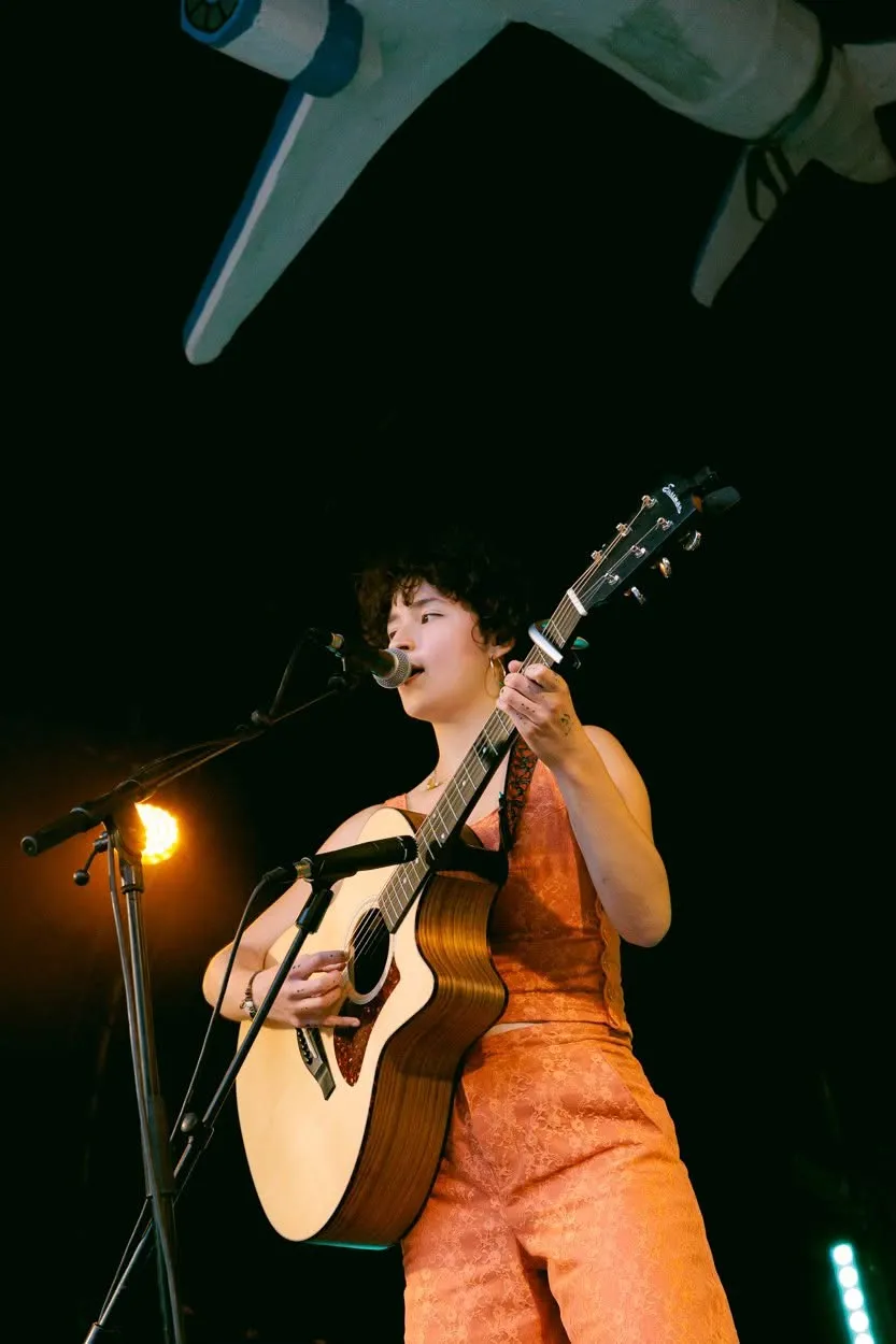 Een zangeres in een oranje outfit speelt akoestische gitaar, onder de vleugel van het vliegtuig.
